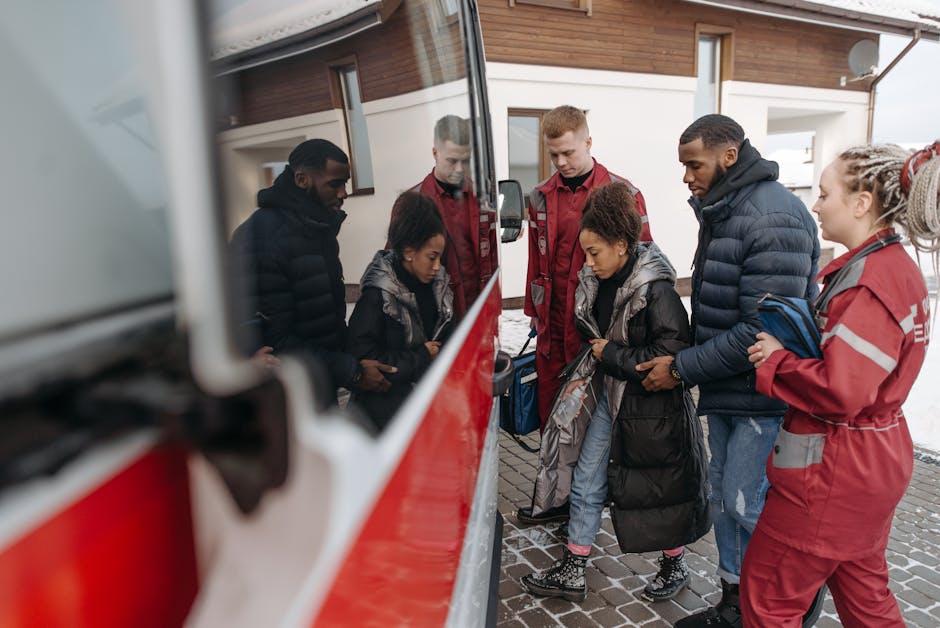 A group of six individuals gathered outdoors in front of a modern residential building with wooden cladding and large windows, observing a red and white waste collection vehicle parked on a paved driv