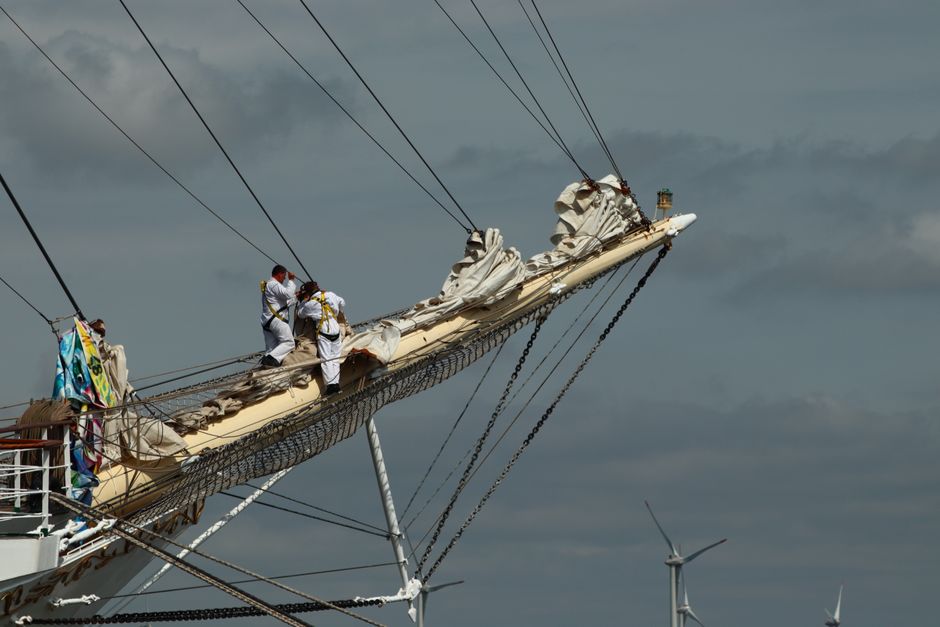 Three workers dressed in white safety uniforms and helmets are seen on the bow of a sailing ship’s wooden mast, which is partially covered in white sails that appear to be furled or folded. The mast