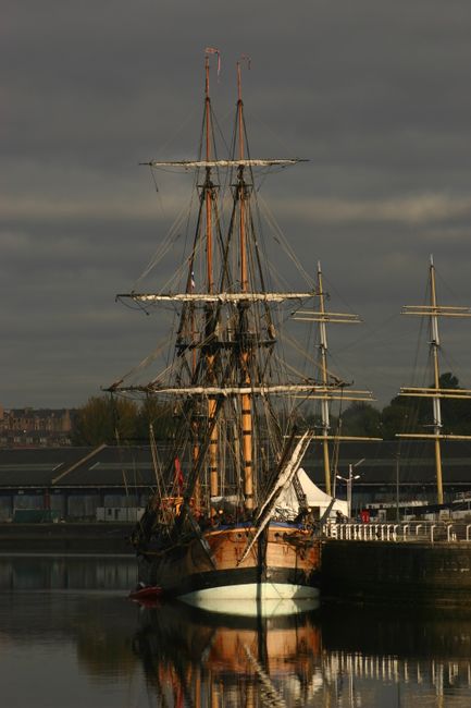 Three workers dressed in white safety uniforms and helmets are seen on the bow of a sailing ship’s wooden mast, which is partially covered in white sails that appear to be furled or folded. The mast