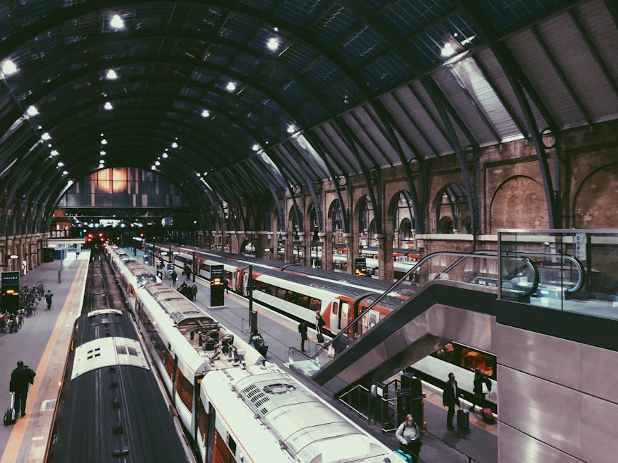 Interior view of a busy train station concourse featuring a large, modern glass and steel structure resembling a twisting tree trunk supporting a domed glass roof. The concourse floor is filled with t
