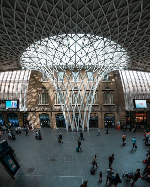 Interior view of a busy train station concourse featuring a large, modern glass and steel structure resembling a twisting tree trunk supporting a domed glass roof. The concourse floor is filled with t