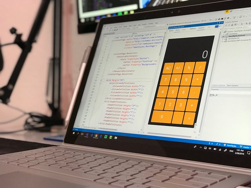 A close-up image of a silver laptop placed on a desk, displaying a software development environment with lines of code on the left side and a large, orange calculator-style interface on the right. The